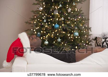 Stock Photo - USA, Illinois, Metamora, Man in Santa hat lying on sofa near Christmas tree. Fotosearch