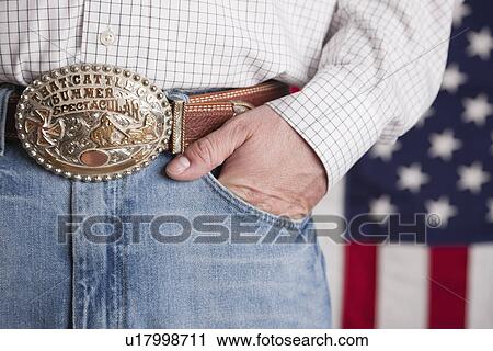 Stock Image - USA, Illinois, Metamora, Man wearing jeans and ornate belt standing in front of American flag, mid section. Fotosearch