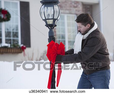 Stock Image - USA, Illinois, Metamora, mid adult man decorating lamp post in front of house with Christmas ribbon. Fotosearch