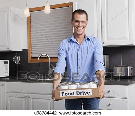 Stock Image - USA, Illinois, Metamora, Portrait of smiling man holding cardboard box with canned food in kitchen. Fotosearch