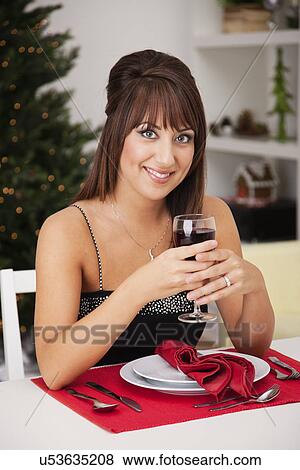 Stock Photo - USA, Illinois, Metamora, Portrait of smiling young woman sitting at table with wineglass, Christmas tree in background. Fotosearch