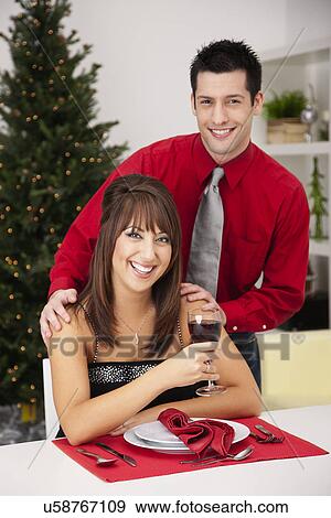 Stock Photo - USA, Illinois, Metamora, Portrait of smiling young Couple at table with wineglass, Christmas tree in background. Fotosearch