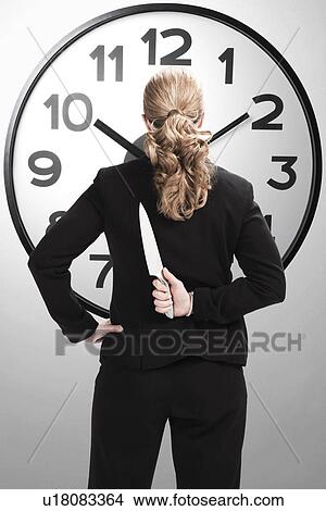 Picture - USA, Illinois, Metamora, Rear view of businesswoman standing if front of large clock, holding kitchen knife. Fotosearch