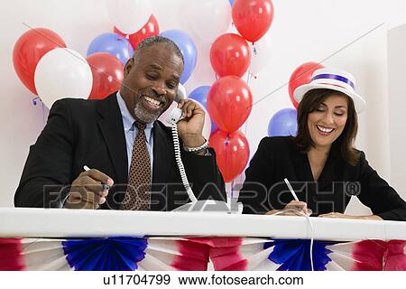 Stock Photo - USA, Illinois, Metamora, Smiling man and woman at polling place table, red and blue balloons in background. Fotosearch