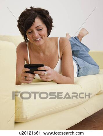 Stock Image - USA, Illinois, Metamora, Smiling young woman lying on sofa with smart phone. Fotosearch