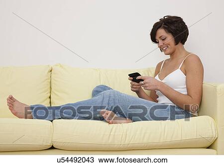 Stock Image - USA, Illinois, Metamora, Smiling young woman sitting on sofa with smart phone. Fotosearch
