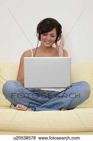 Stock Photo - USA, Illinois, Metamora, Smiling young woman with headphones sitting on sofa and using laptop. Fotosearch