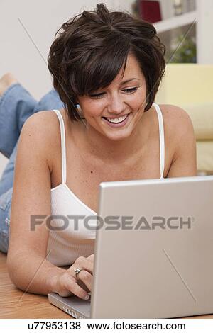 Stock Photo - USA, Illinois, Metamora, Smiling young woman lying on floor with laptop. Fotosearch