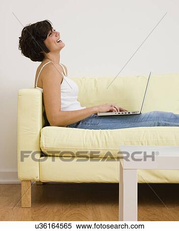 Stock Photography - USA, Illinois, Metamora, Smiling young woman sitting on sofa with laptop. Fotosearch