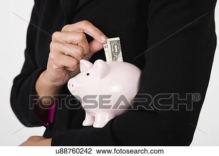 Stock Image - USA, Illinois, Metamora, Woman putting one dollar banknote into piggy bank, mid section. Fotosearch