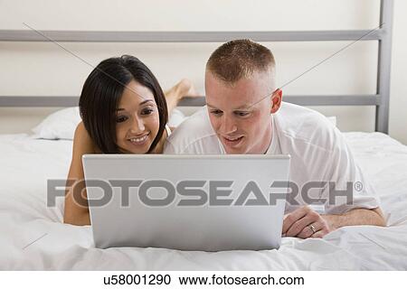Stock Image - USA, Illinois, Metamora, Young couple lying in bed and using laptop. Fotosearch