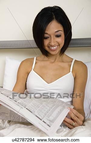 Picture - USA, Illinois, Metamora, Young woman sitting in bed and reading newspaper. Fotosearch