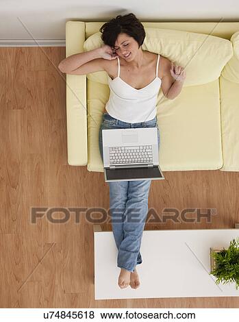 Stock Photo - USA, Illinois, Metamora, Young woman sitting on sofa with laptop. Fotosearch
