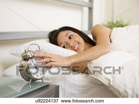 Stock Photo - USA, Illinois, Metamora, Young woman turning off alarm clock in bed. Fotosearch