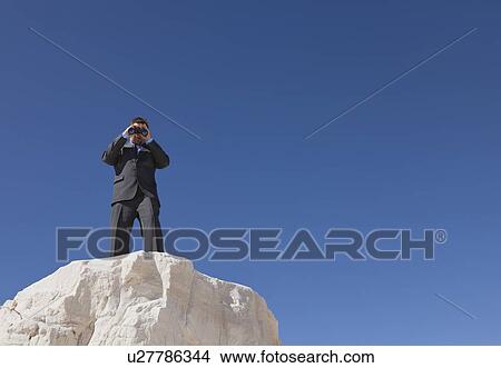 Picture - USA, New Mexico, Whitesands, Businessman on rock looking through binoculars. Fotosearch