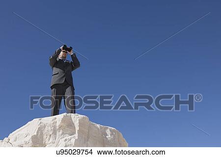 Picture - USA, New Mexico, Whitesands, Businessman on rock looking through binoculars. Fotosearch