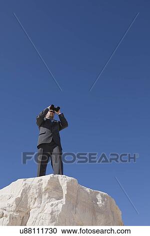 Stock Image - USA, New Mexico, Whitesands, Businessman on rock looking through binoculars. Fotosearch