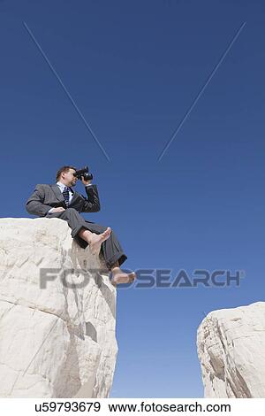 Stock Photo - USA, New Mexico, Whitesands, Businessman looking through binoculars and sitting at rock. Fotosearch