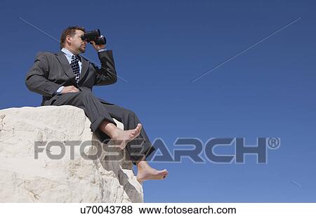 Stock Photo - USA, New Mexico, Whitesands, Businessman looking through binoculars and sitting at rock. Fotosearch