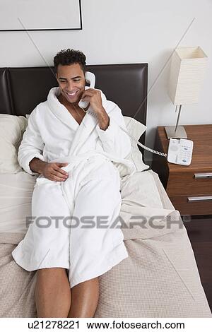Stock Image - USA, New York City, man sitting on bed and talking on phone. Fotosearch