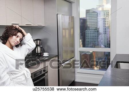 Stock Photo - USA, New York City, woman standing in kitchen with hand in hair. Fotosearch