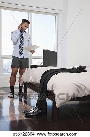 Picture - USA, New York, New York City, man looking at document and talking on mobile phone in bedroom. Fotosearch