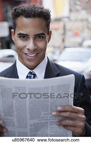 Stock Image - USA, New York, New York City, portrait of smiling businessman holding newspaper on street. Fotosearch