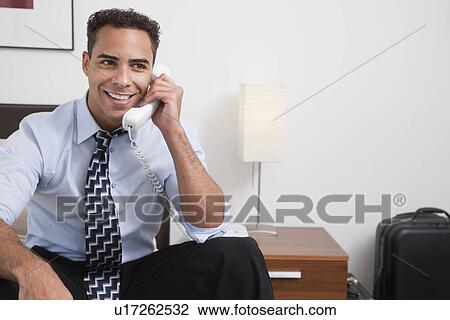 Stock Image - USA, New York, New York City, smiling young man sitting talking on mobile phone in hotel room. Fotosearch