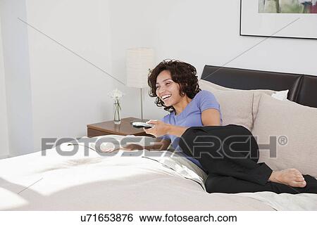 Stock Photograph - USA, New York, New York City, smiling young woman lying on bed and watching TV in hotel room. Fotosearch