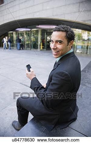 Stock Image - USA, New York, New York City, Businessman with cell phone sitting in front of store. Fotosearch