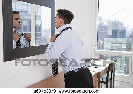 Stock Photo - USA, New York, New York City, young man tying tie in front of mirror in hotel room. Fotosearch