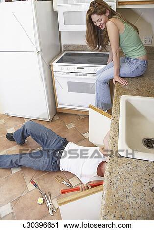 Stock Image - Young woman watching plumber fixing kitchen sink. Fotosearch