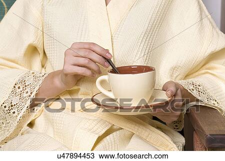 Stock Image - Young woman wearing bathrobe holding tea cup, mid section. Fotosearch