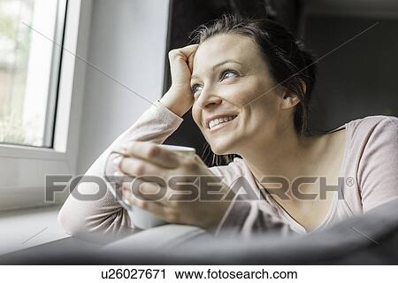 Stock Image - Mid adult woman looking out of window with coffee. Fotosearch