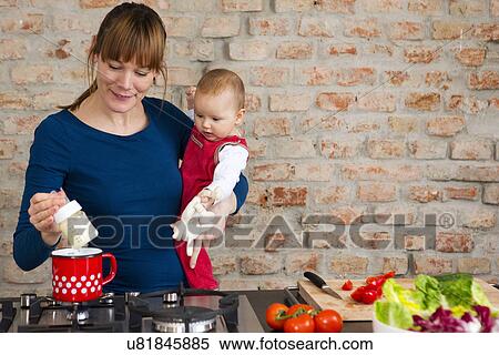 Stock Photography - Mother carrying baby daughter whilst preparing lunch. Fotosearch