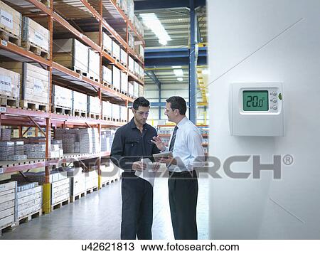 Stock Image - Office manager and worker discuss energy use near thermostat in factory. Fotosearch