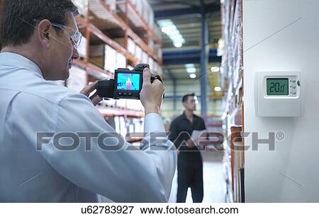 Stock Photo - Office worker taking thermal image with camera in factory next to thermostat. Fotosearch