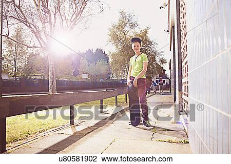 Stock Image - Portrait of boy standing on pavement holding skateboard. Fotosearch