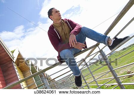 Stock Image - Portrait of young man sitting on beach hut fence. Fotosearch