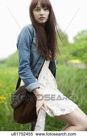 Stock Photography - Portrait of young woman sitting on fence in field. Fotosearch