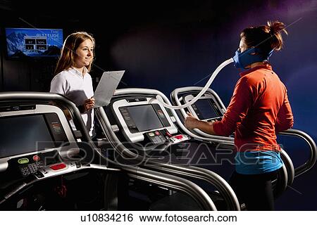 Stock Photograph - Trainer monitoring mid adult woman with face mask on gym treadmill in altitude centre. Fotosearch