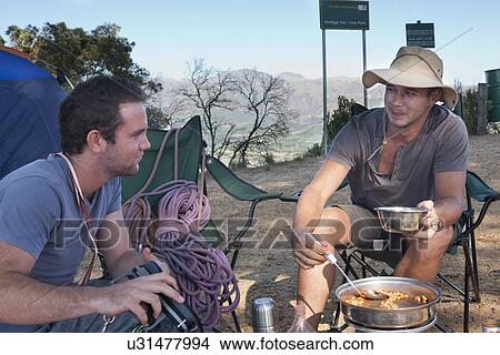 Picture - Two young male campers chatting and preparing meal. Fotosearch