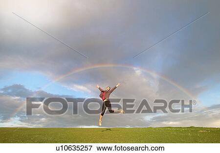 Stock Photo - Young female hiker jumping mid air below rainbow. Fotosearch