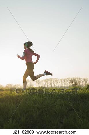 Stock Photo - Young female runner listening to headphones whilst running. Fotosearch