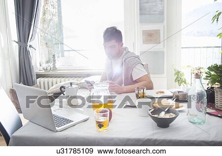 Stock Photo - Young man having breakfast and using laptop. Fotosearch