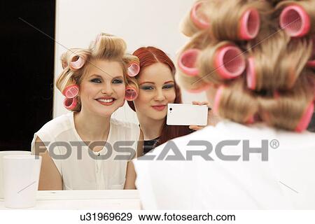 Stock Photo - Young woman in curlers and friend taking selfie. Fotosearch