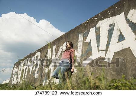 Stock Image - Young woman leaning against wall with graffiti. Fotosearch
