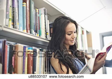 Stock Photo - Young woman reading book by bookshelf. Fotosearch
