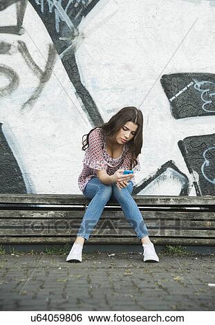 Stock Photograph - Young woman sitting on bench using cell phone. Fotosearch