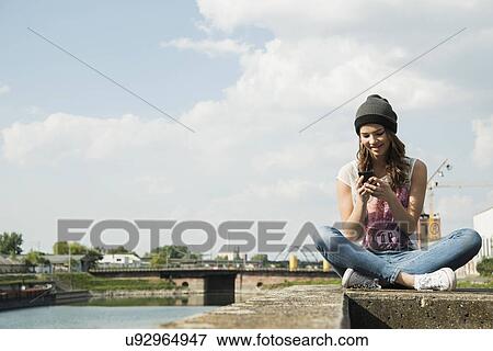 Young woman sitting on wall using cell phone View Large Photo Image Stock Photo - Young woman sitting on wall using cell phone. Fotosearch